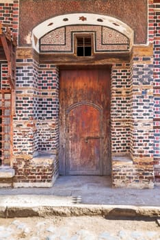Wooden grunge decorated arched entrance gate with inner small wooden door on wall with black and red bricks with white seam
