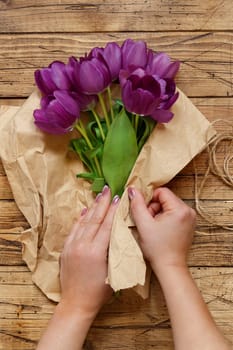 Hands wrapping purple tulips bouquet in craft paper overwooden table top view. Rustic valentines gift