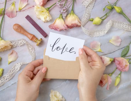 Hands with Card LOVE inside envelope near by pink flowers, wax seal and ribbons on a marble table top view. Romantic scene with handwritten card flat lay. Valentines concept