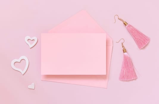 Pink paper card and envelope, hearts and tassel earrings on pink top view. Minimal scene with blank card mockup. Valentines day, educational and girly concept, space for text.