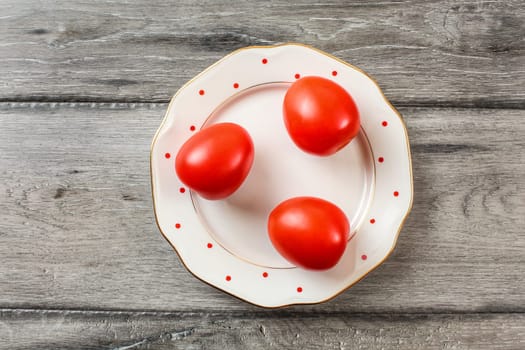 Three whole tomatoes on white plate with small red dots and golden rim, gray wooden desk, tabletop view.