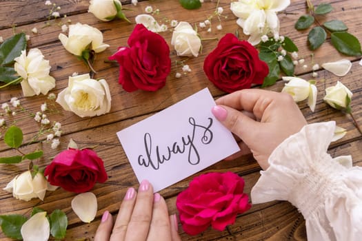 Hands with handwritten card ALWAYS surrounded by red and cream roses close up on a wooden table. Femminine romantic declaration of love near flowers, Valentines day concept