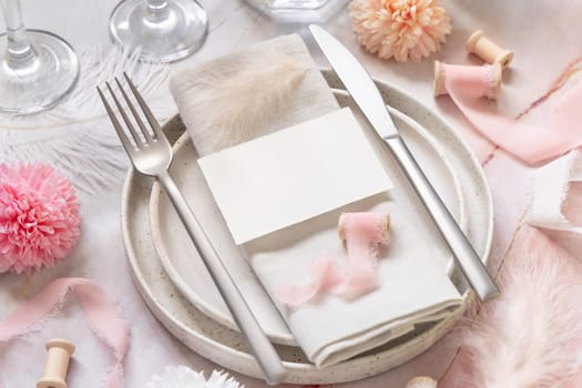 Wedding Table place with a paper card over napkin near flowers, silk ribbons and feathers closeup. Romantic template with blank card on a plate flat lay. Vertical card mockup, copy space