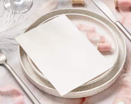 Wedding Table place with a blank card near flowers, silk ribbons and feathers, closeup. Romantic scene with vertical paper card mockup on a plate on a marble table
