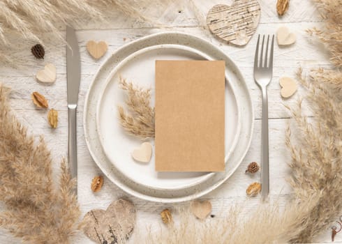 Wedding table place with blank card near dried plants, palm leaves, pampas grass and hearts top view. Boho scene with vertical kraft paper card flat lay. Greeting, Invitation or valentines mockup
