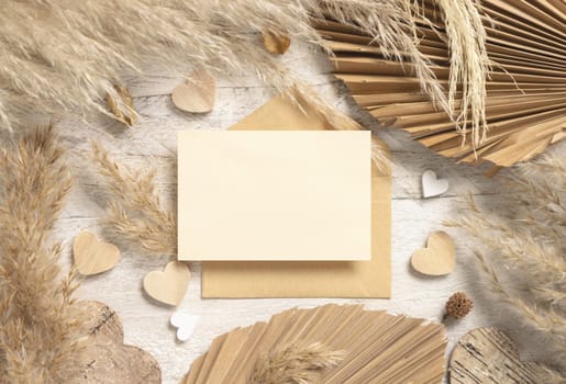 Blank card and envelope on white wooden table near dried pampas grass and hearts top view. Boho scene with horizontal card, flat lay. Bohemian Valentines mockup