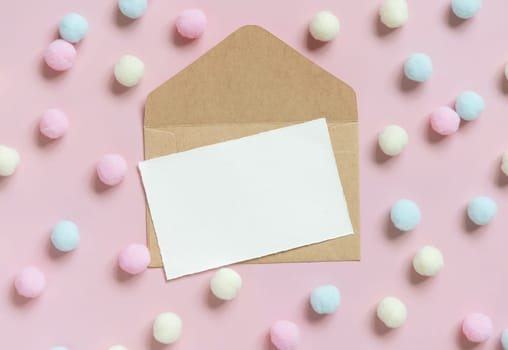 Blank card on envelope near pastel pom-poms on light pink top view. Romantic scene with blank horizontal card with place for text, flat lay. Valentines, Spring or girlish concept