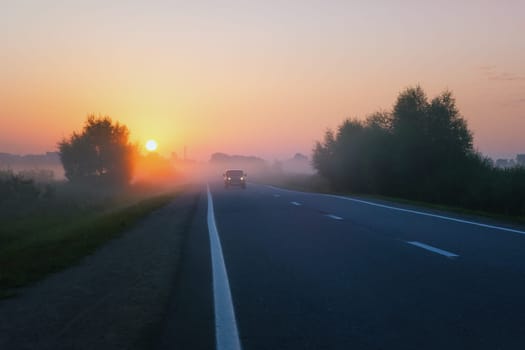 Misty golden sunrise above an suburban asphalt road in the countryside. Car with headlights in the distance drives out of the fog in the early morning at dawn.