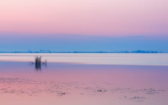 Quiet, gently pink lilac dawn by the lake. Clear sky over calm water surface and misty blue horizon. Tranquil lakeshore background with copy space. Russia, Lake Nero.