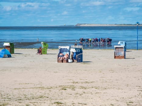Sunny beach scene with beach chairs on the sandy beach in the foreground and a guided group at the start of a mudflat hike and a view of the island of Spiekeroog in the background under a blue sky with individual clouds.
