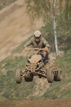 Quad racer is high jumping. The quad bike and rider are very muddy.Potential trademarks are removed and face of the racer is unidentifiable.