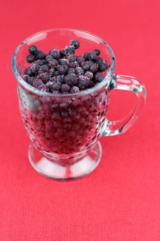 Dark wild frozen blueberries with some ice crystals thawing in a clear glass coffee cup on a red background. Close-up of purple frozen wild blueberries being thawed in a clear glass cup with handle.