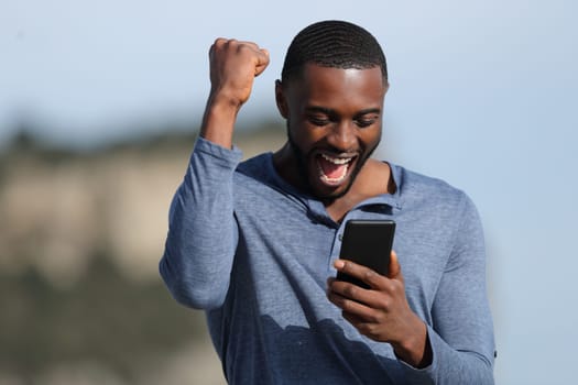 Excited man with black skin celebrating good news checking smart phone in the mountain