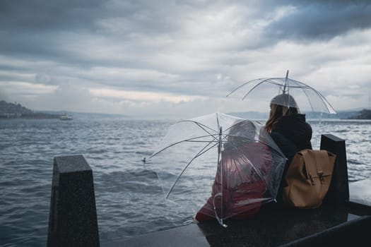 rear view closeup of mother and daughter holding transparent umbrellas sitting on shore and watching the sea on a rainy day with storm clouds above
