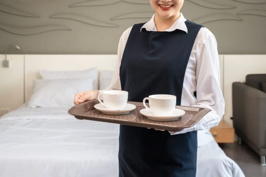 Female Chambermaid holding tray with two cups of coffee and standing next to white bed in guest room. Hotel room service concept.