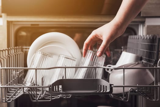 Female hand loading dished, empty out or unloading dishwasher with utensils. Kitchen appliances, lifestyle view. Woman puts a plate in the dishwasher or takes from it. Housewife does her housework