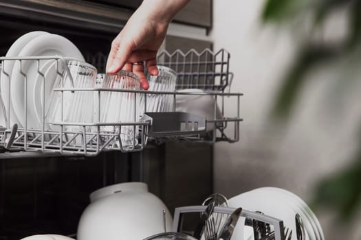 Close-up of female hand loading dished to, empty out or unloading from open automatic stainless built-in dishwasher machine with clean utensils inside in modern home kitchen. Household domestic life