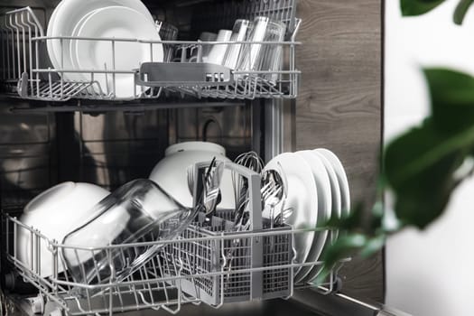 Front view of open automatic stainless steel built-in fully integrated top control dishwasher range machine with clean utensils, cutlery, glasses, dishes, plates inside in modern home kitchen