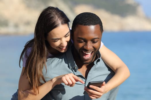 Front view portrait of an interracial couple checking phone enjoying together on the beach on summer vacation