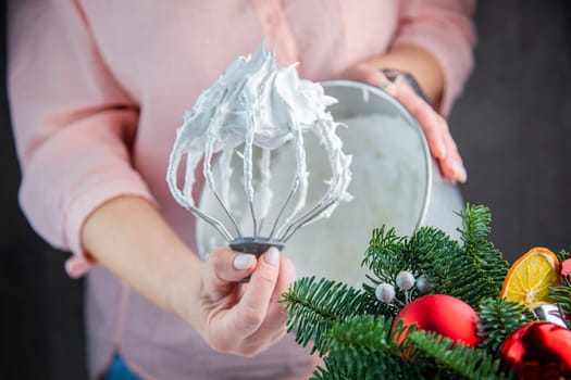 The whisk of an electric mixer in a white mass of meringue with standing peaks is held in the hand of a pastry chef against the background of an iron bowl, the branches of a Christmas tree and New Year's toys are visible from the side. High quality photo
