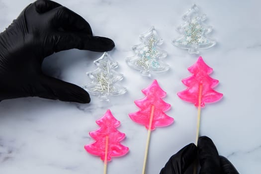 Three pink lollipops in the form of Christmas trees lie on white marble, above them are white Christmas trees - lollipops with powder, the extreme ones hold hands in black gloves. High quality photo