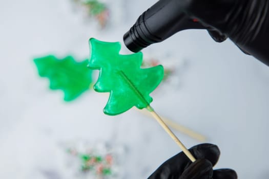 A green lollipop in the form of a Christmas tree is held in the hands of black gloves and burned on fire from a lighter, in the process of making a lollipop. High quality photo