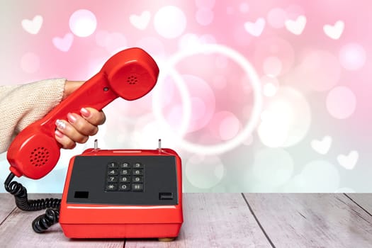 A feminine elegant beautifully manicured woman's hand holding an old red telephone handset over an abstract blurry light pink Valentine's Day or wedding background. Copy Space. Communication background.