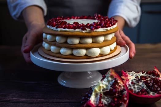 The cake stands on a white stand, decorated with cream and pomegranate on top, it is held by the hands of a pastry chef, there is a pomegranate next to it and there are flowers on a dark background. High quality photo