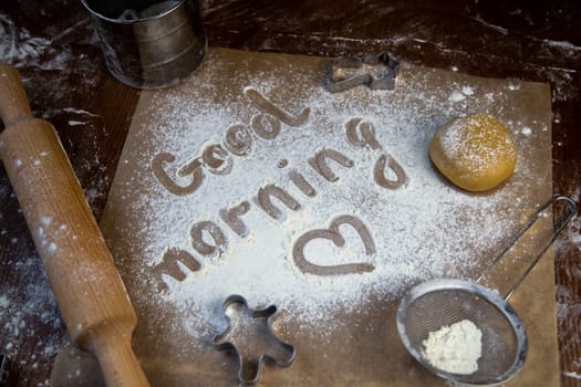On the baking paper on the flour is written Good morning and heart, against the background of dough, rolling pins and cookie cutters. High quality photo