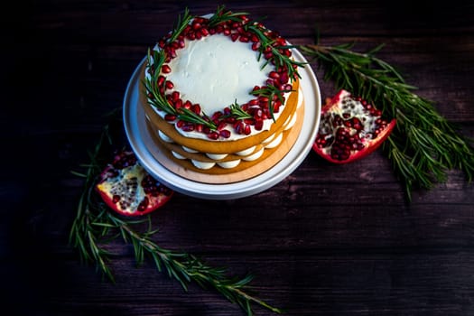 Cake with white cream and honey brown cakes lies on a white stand, decorated with pomegranate and rosemary, against a dark tree. High quality photo