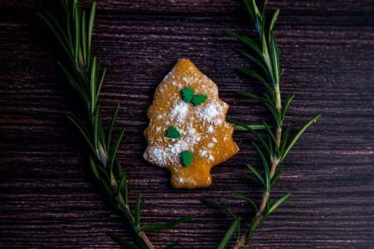 Cookies in the form of a Christmas tree, sprinkled with powdered sugar and small Christmas trees on top, lies on a dark wooden background surrounded by grained rosemary. High quality photo