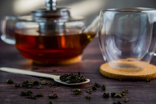 A wooden spoon with tea lies against a dark wood background, a mug and a teapot are visible from behind, a photo in dark colors. High quality photo