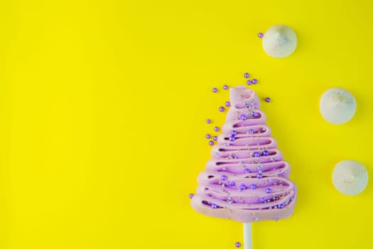 Pale purple Christmas tree made of meringue on a stick, decorated with purple and silver balls on a bright yellow background, purple powder balls and white snowdrifts are scattered near the tree. High quality photo
