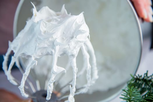 A whisk from a metal mixer in a white mass of bizet on the background of a bowl, the hands of a pastry chef and the branches of a Christmas tree are visible. High quality photo