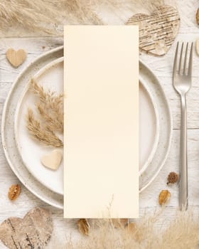 Wedding table place with blank menu card near dried plants, palm leaves, pampas grass and hearts top view. Boho scene with vertical paper card flat lay. Valentines mockup