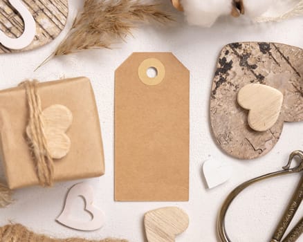 Kraft paper gift tag between Valentines present, wooden hearts and dried flowers top view. Spring scene with vertical blank label. Rustic, cottage chic or bohemian mockup, copy space