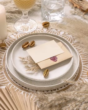 Wedding table place on bohemian placemat with horizontal blank card close up. Boho wedding card mockup. Tropical beige template with dried plants, palm leaves and pampas grass
