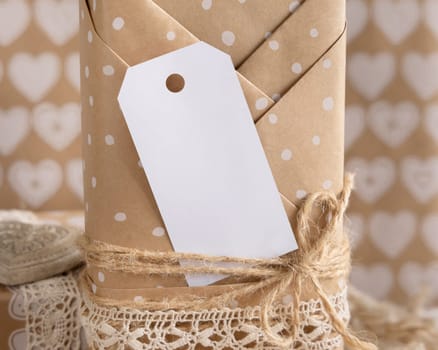 Valentines kraft paper wrapped present with vertical gift tag and hearts close up. Spring scene with blank label. Rustic, cottage chic or bohemian mockup, copy space