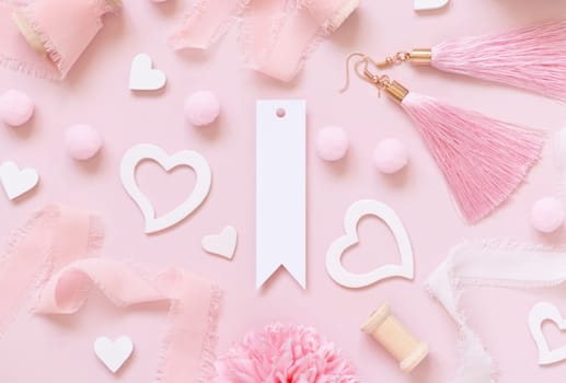 Blank vertical gift tag, hearts, flowers, silk ribbons and tassel earrings on pink top view. Romantic scene with blank label mockup flat lay. Valentines day, wedding and girly concept.