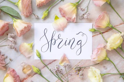 Card with the handwritten text SPRING surrounded by pink flowers, earrings, petals and parfume flacon top view on a marble table. Romantic concept