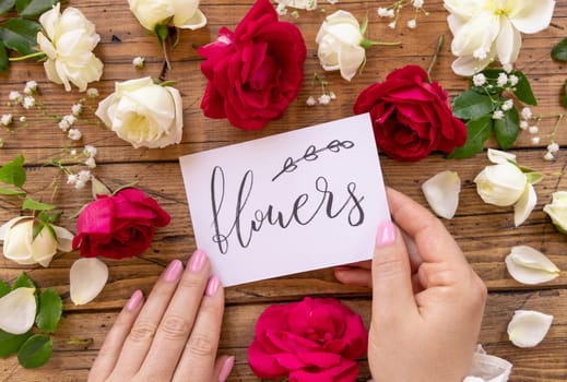 Hands with handwritten card FLOWERS surrounded by red and cream roses close up on a wooden table. Femminine romantic declaration of love near flowers, Valentines day concept