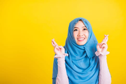 Asian Muslim Arab, Portrait of happy Asian beautiful young woman Islamic religious wear veil hijab she smiling and holding fingers crossed for good luck, studio shot isolated on yellow background