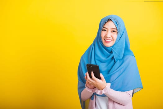 Asian Muslim Arab, Portrait of happy beautiful young woman Islam religious wear veil hijab funny smile she using hold mobile smart phone reading social media isolated on yellow background