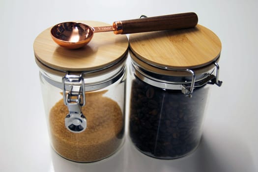 Close up of Two isolated glass and wood jars containing coffee and cane sugar with a copper and wood table spoon