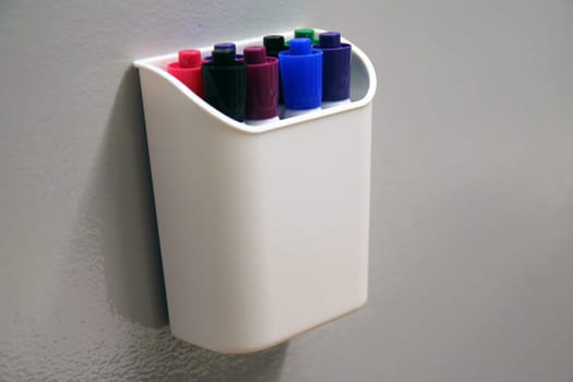 The marking pens in their container hanging on a erasable grey wall of an office