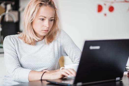Female freelancer in her casual home clothing working remotly from her dining table in the morning. Home kitchen in the background