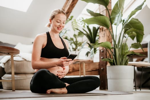 Pregnancy and technoloy supported healthy lifestyle concept. Cheerful happy pregnant woman using smart phone application while exercising on yoga mat on living room floor at home