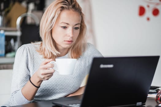 Female freelancer in her casual home clothing working remotly from her dining table in the morning. Home kitchen in the background