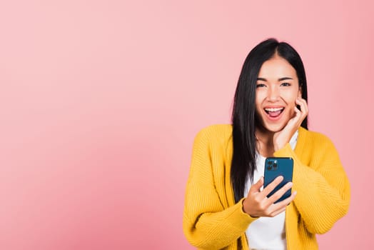 Happy Asian portrait beautiful cute young woman teen smiling excited using smart mobile phone studio shot isolated on pink background, Thai female surprised making winner gesture on smartphone