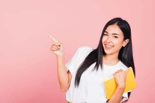 Portrait of happy Asian beautiful young woman confident smiling holding orange book open pointing finger to side copy space, studio shot isolated on pink background, education concept